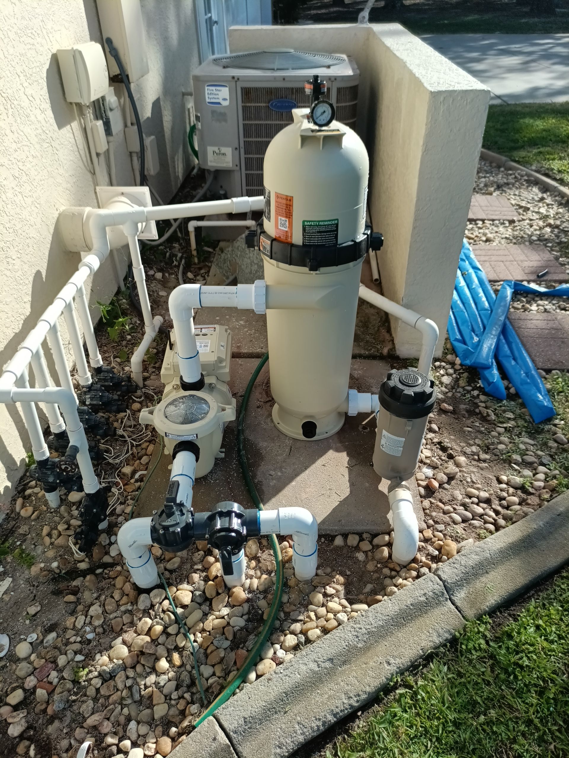 Pool filter system next to a building, with white pipes, beige filter tank, and blue hoses.