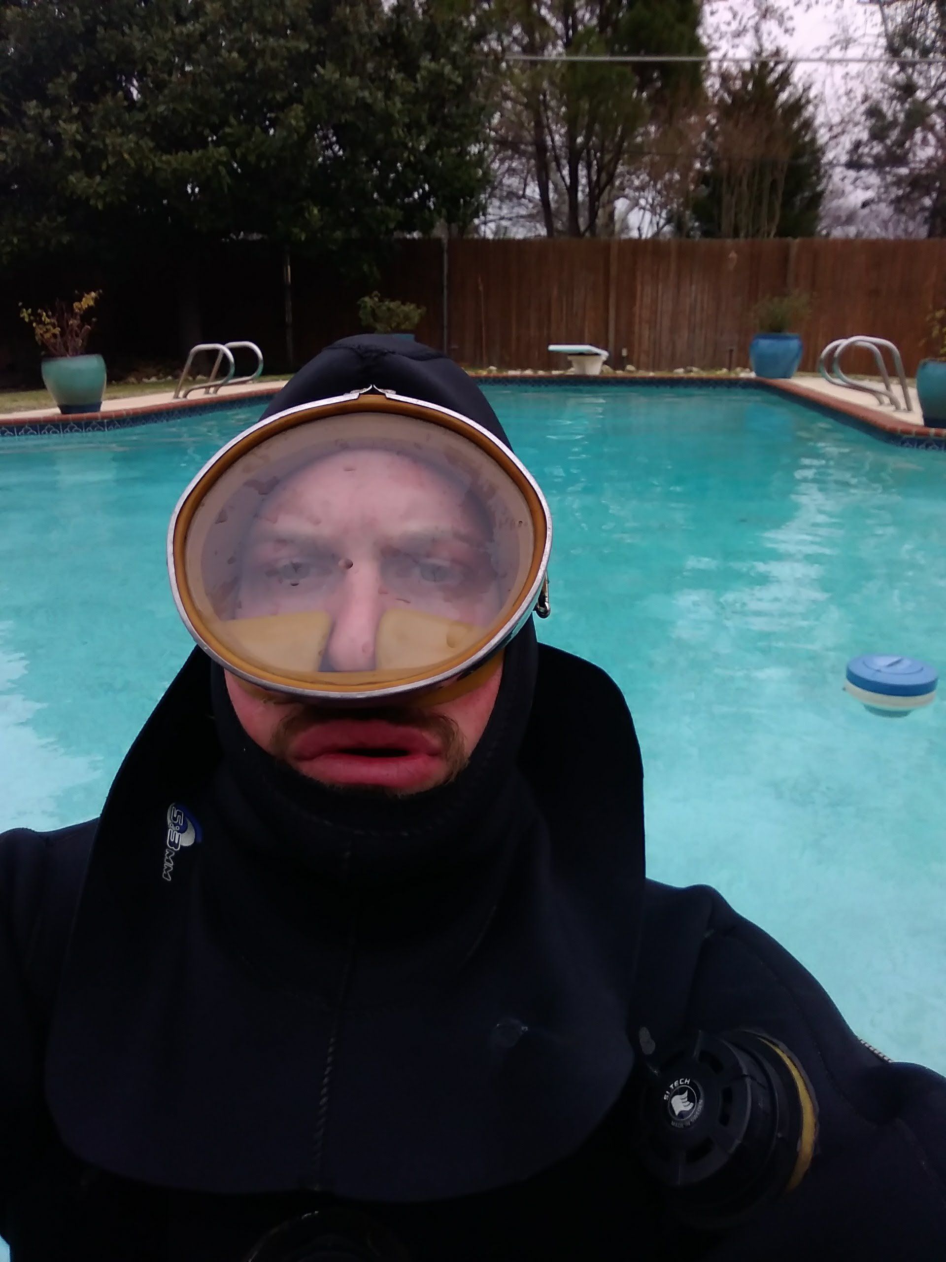 Person in a black diving suit and large goggles, standing in a blue swimming pool.