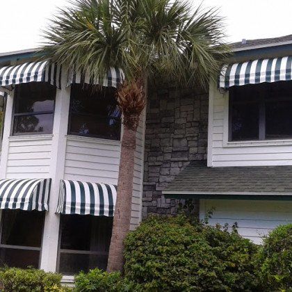 Two-story house with white siding, stone accents, green striped awnings over the windows, and a tall palm tree in front.