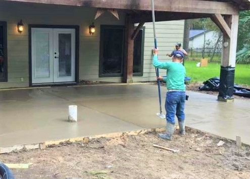 Man smoothing concrete patio with a long tool, near a house with an overhang.