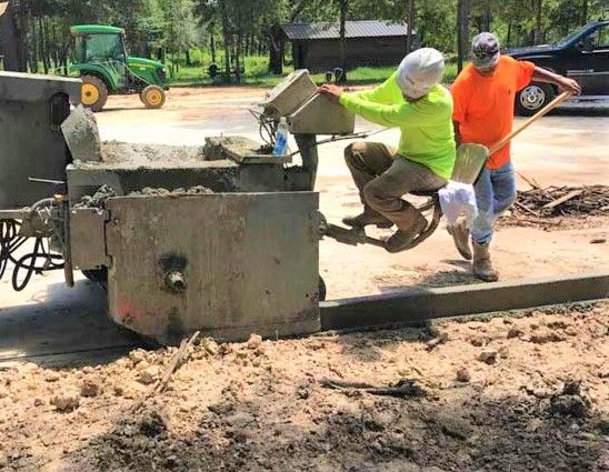 Two workers pour concrete from a mixer into a form on a construction site. One holds the mixer, the other helps.