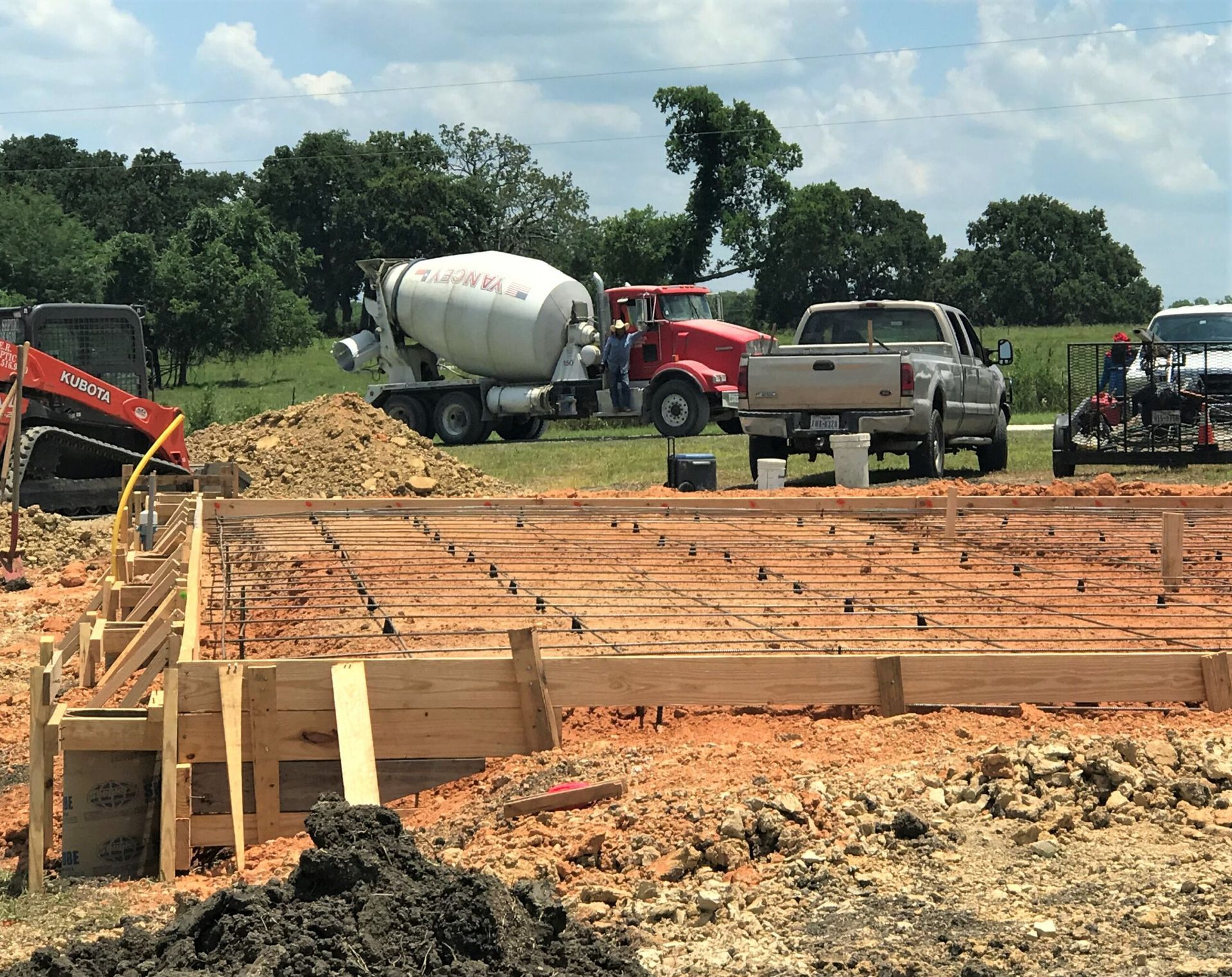 Construction site: concrete forms, rebar, cement truck, and pickup trucks on a dirt foundation, under a sunny sky.