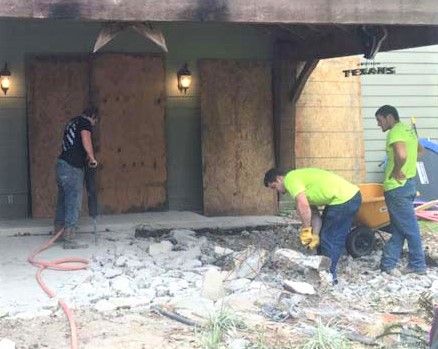 Three workers break concrete in front of a building; plywood covers the doorway.