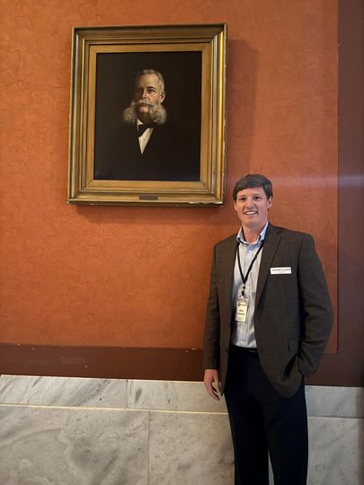 A man in a suit stands in front of a framed portrait of a man