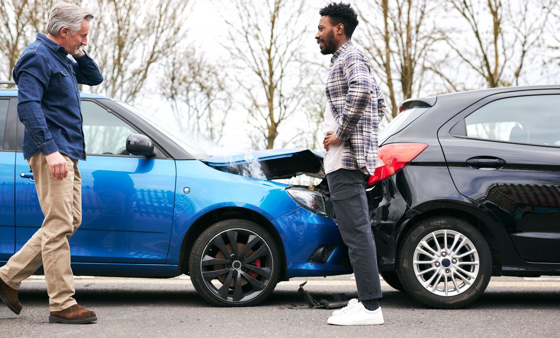 Two men are standing next to each other in front of a car accident.