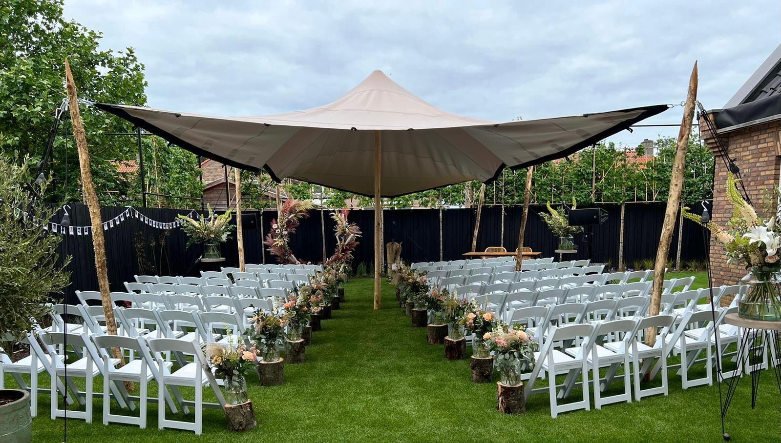 De huwelijksceremonie is ingericht met witte stoelen, bloemversieringen langs het gangpad en een beige baldakijn op een grasveld.
