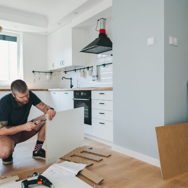 Man assembling furniture in a white kitchen with wood cabinets, using tools.