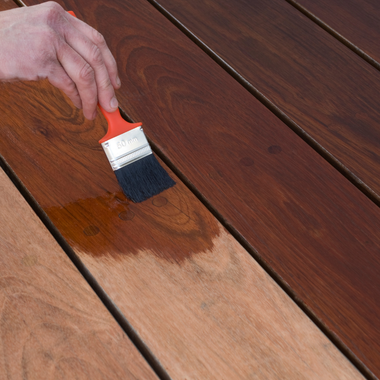 Hand applying wood stain to wooden planks with a paintbrush; some planks stained, others unstained.