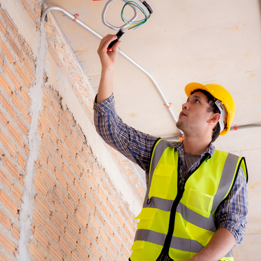 Construction worker in yellow helmet and vest inspecting wiring on a brick wall.