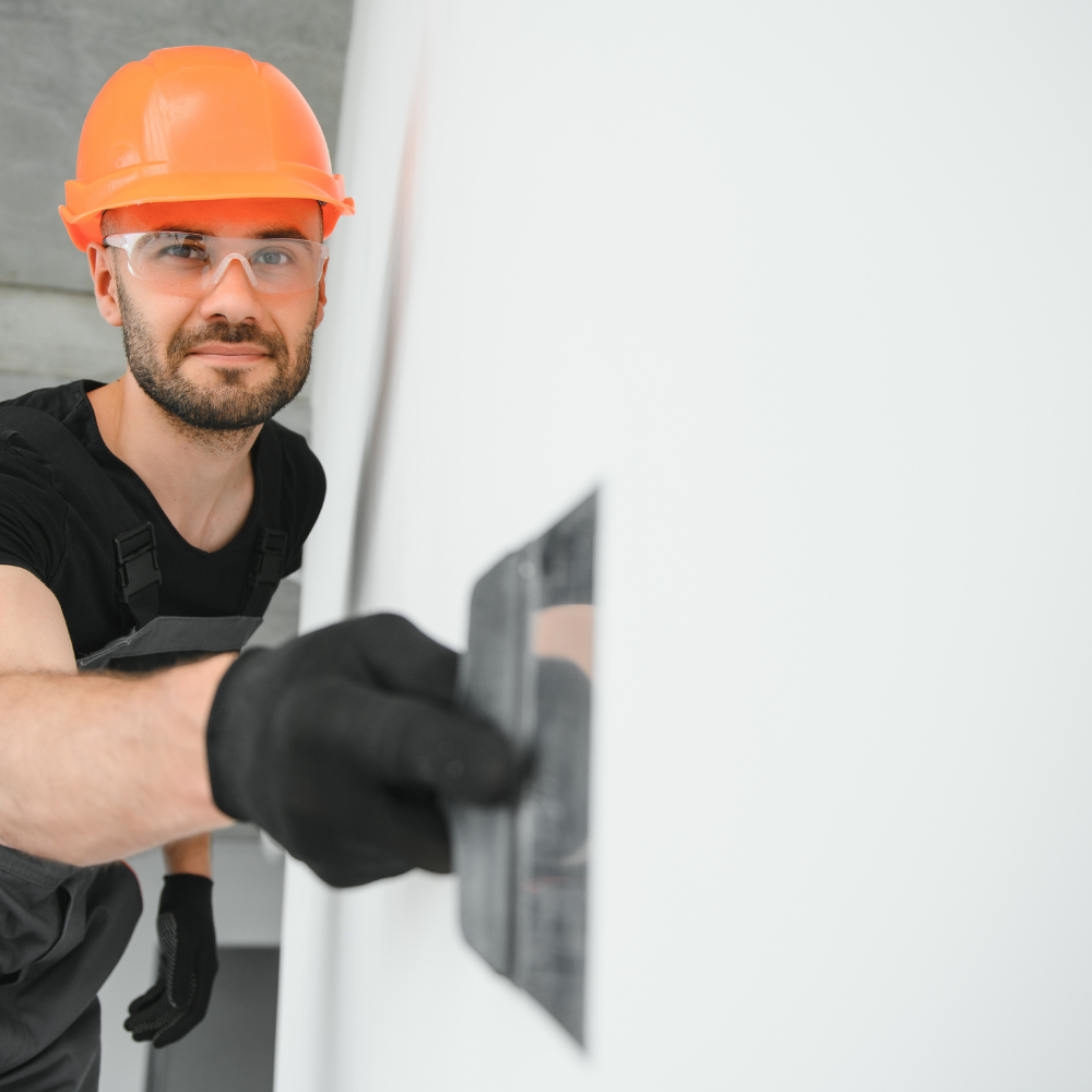 Construction worker in orange hard hat and safety glasses, smoothing plaster on a white wall.