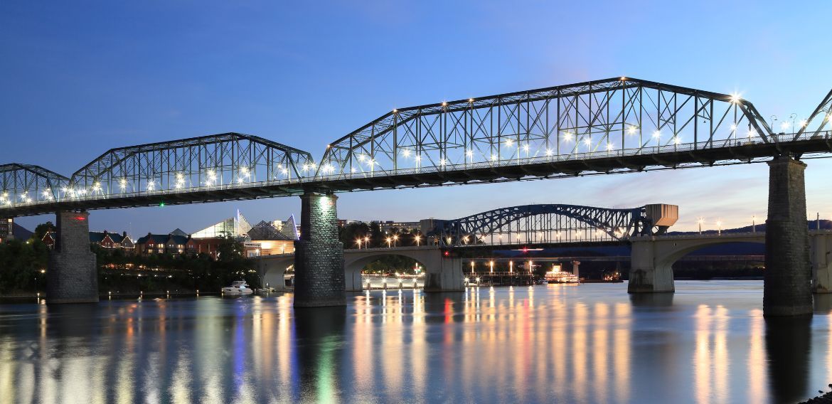A brightly lit bridge reflects in the water at dusk.