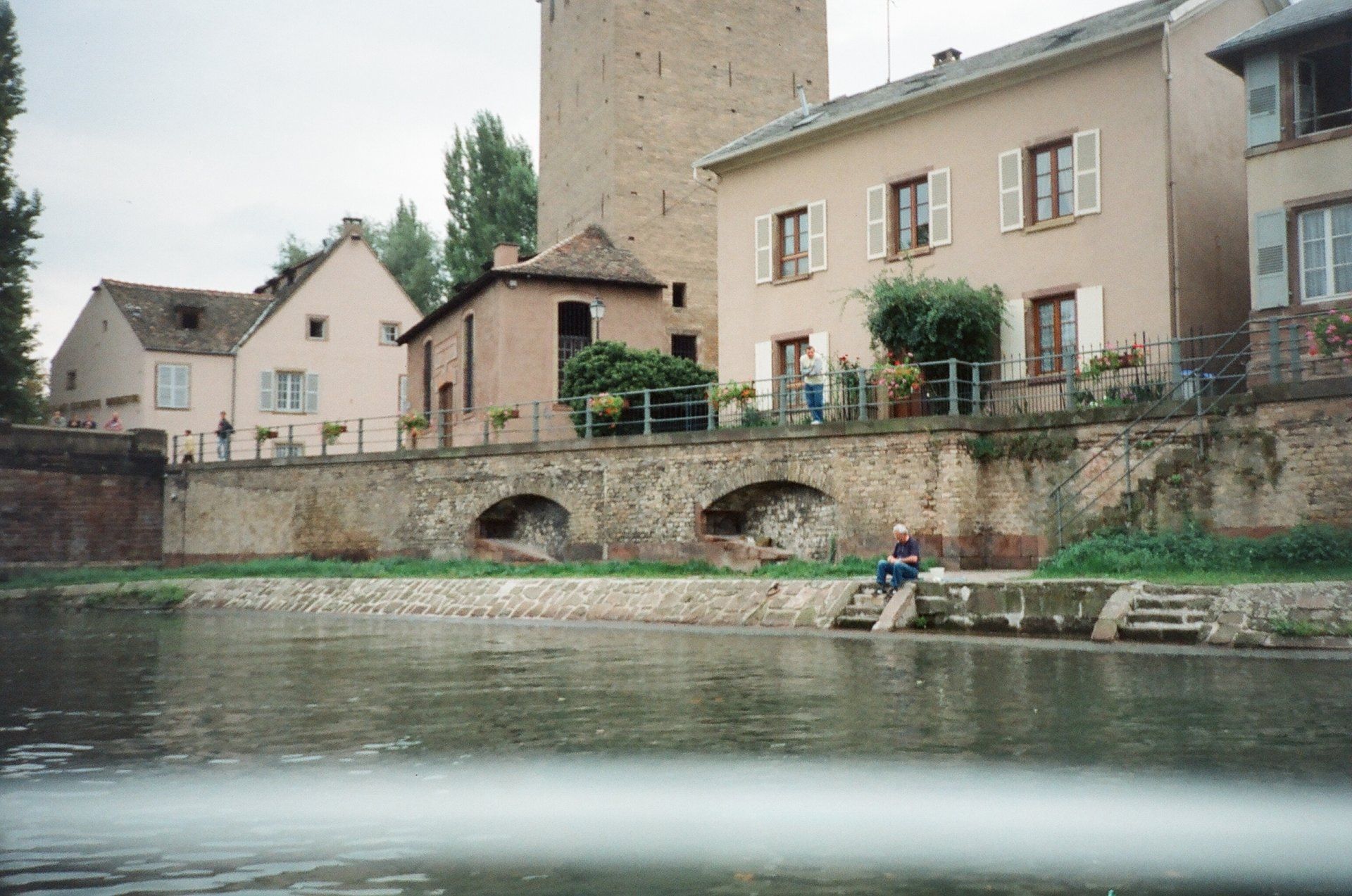 A man sits on a dock next to a river with buildings in the background