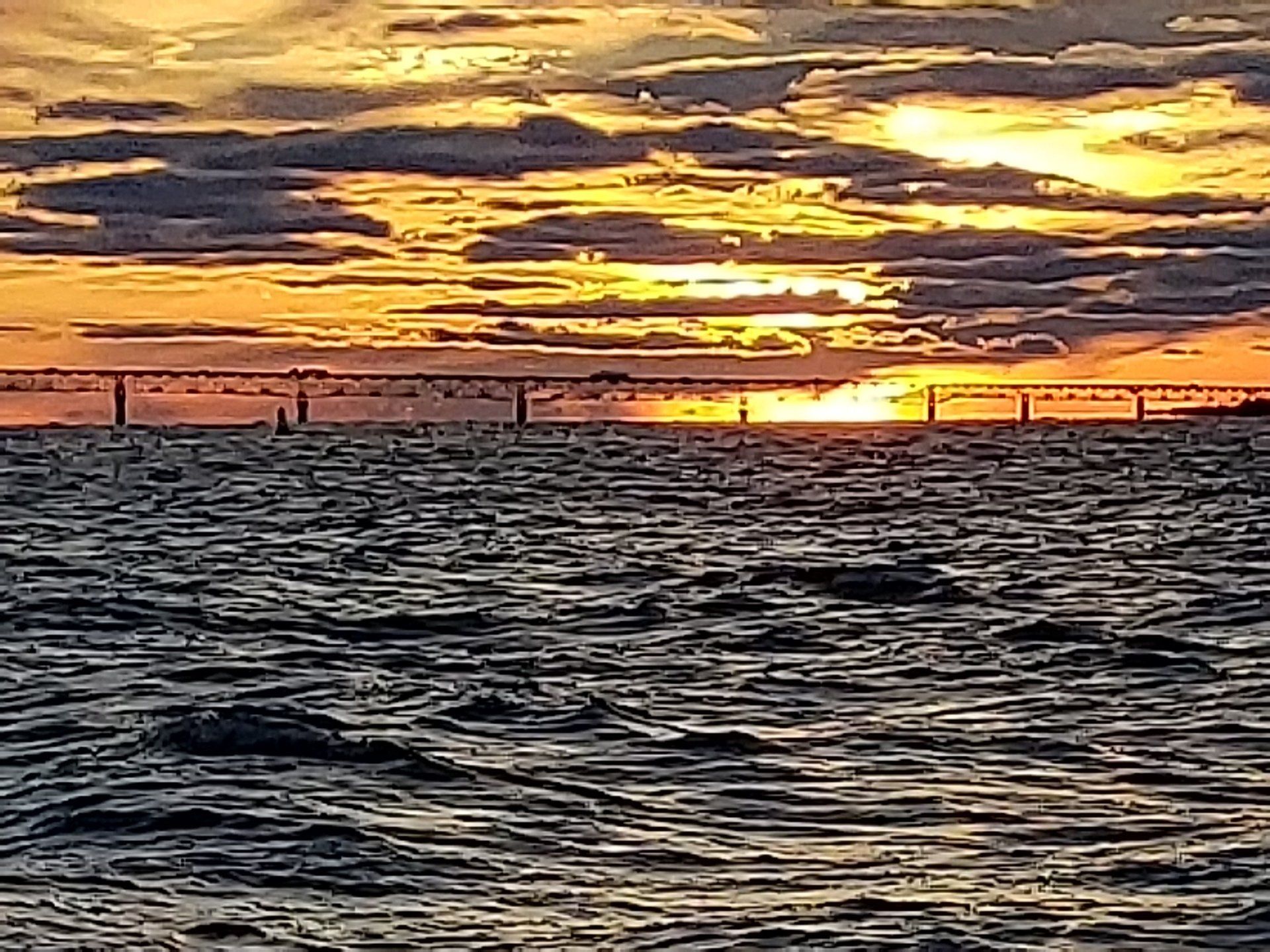 The sun is setting over the ocean with a bridge in the background.