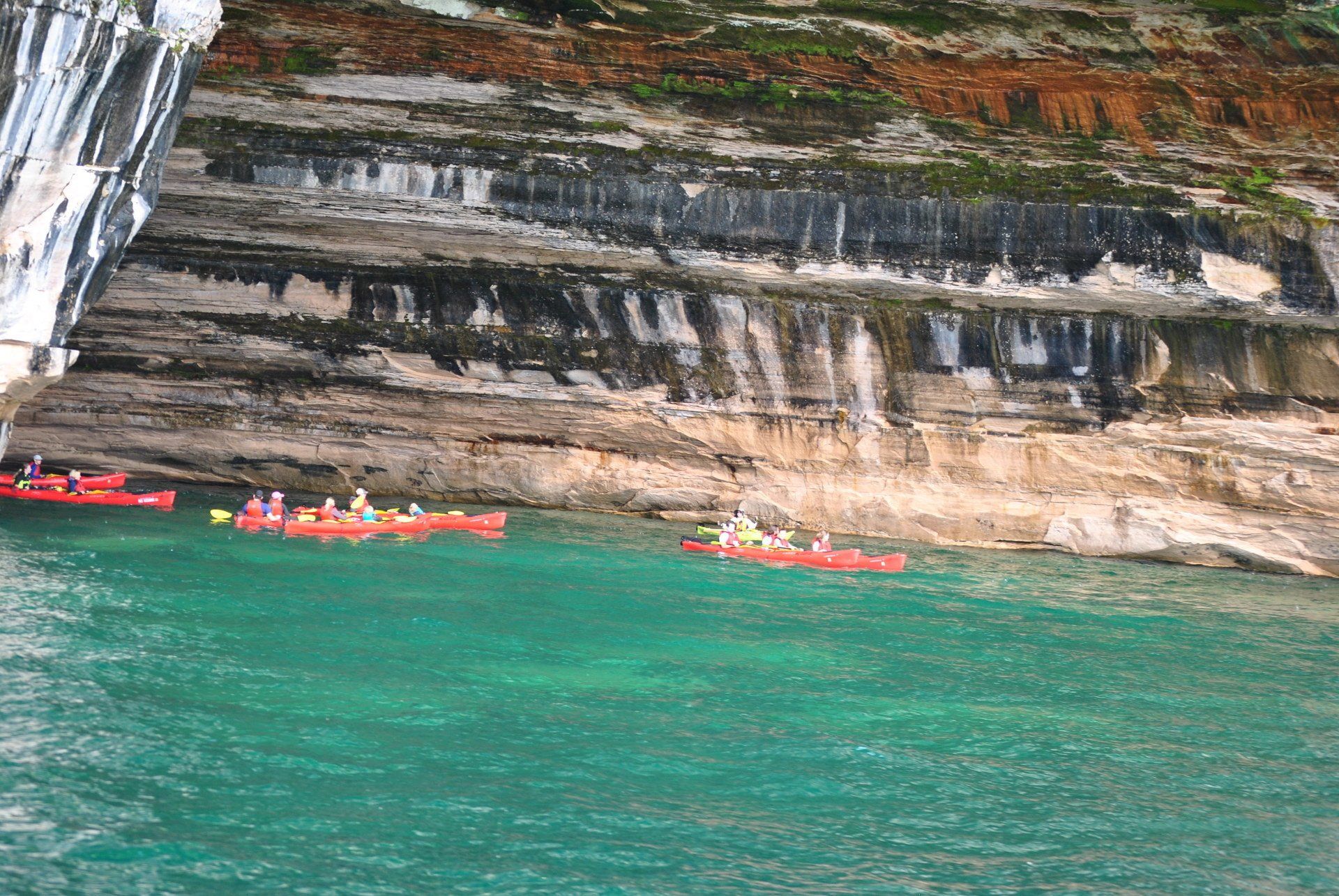 A group of people are in kayaks in the water near a cliff.