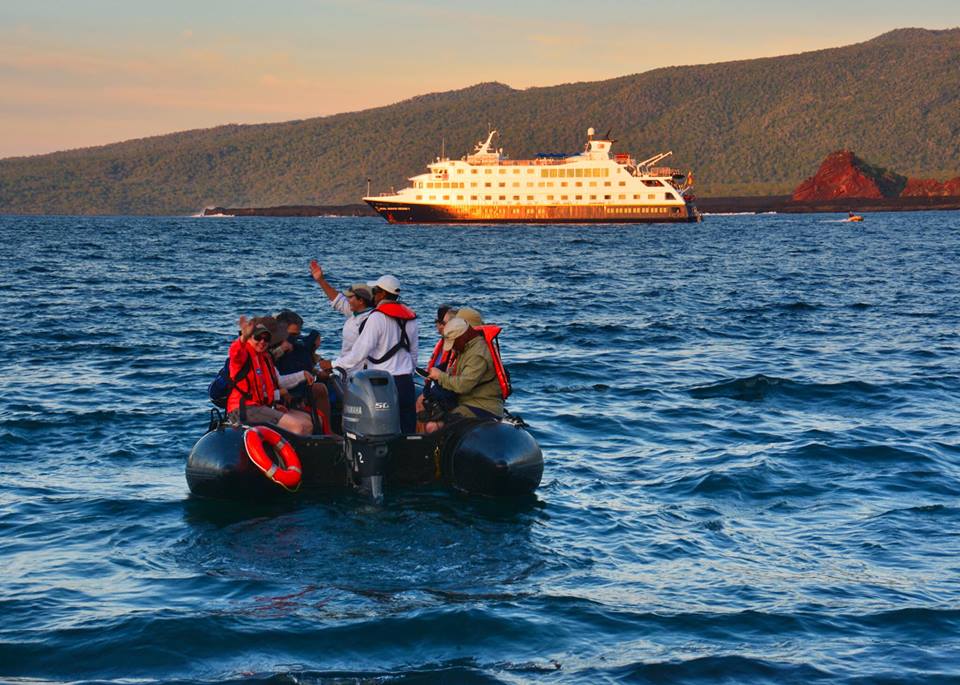 A group of people in a boat in the ocean with a cruise ship in the background.