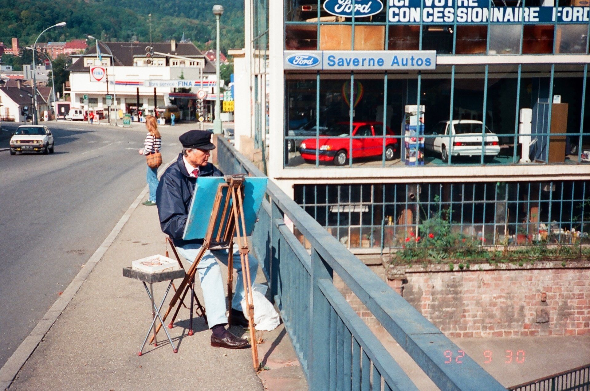 A man sits on a bridge painting in front of a ford dealership