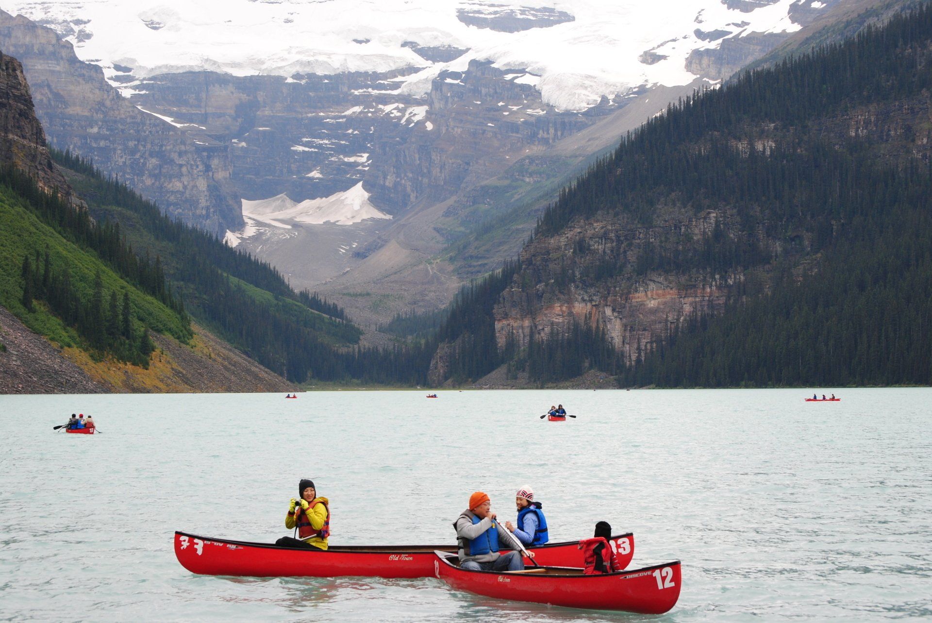 A group of people are in red canoes on a lake with mountains in the background.