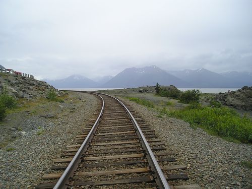 A train track with mountains in the background and a lake in the foreground