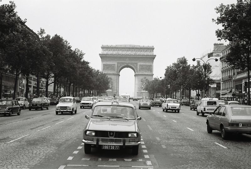 A black and white photo of cars driving down a street with a triumphal arch in the background