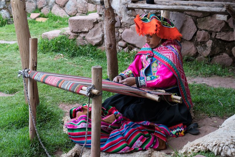 A woman is sitting on the grass using a loom to weave a blanket.