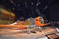 A small orange airplane is on display in a museum.