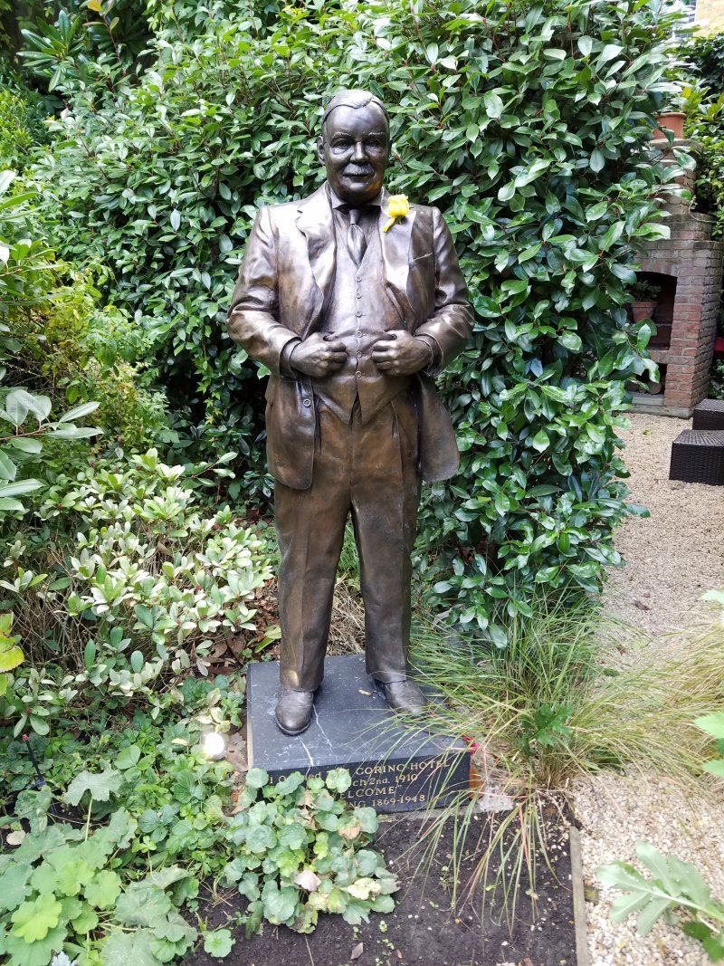 A bronze statue of a man in a suit and bow tie is standing in a garden.