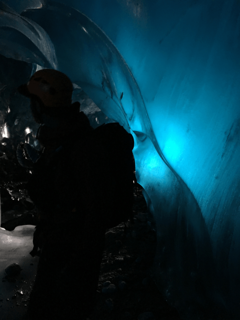 A person is standing in an ice cave with a blue light shining through the ice.