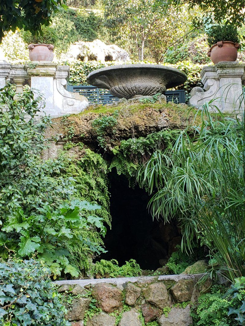 A cave surrounded by trees and plants with a fountain in the background.