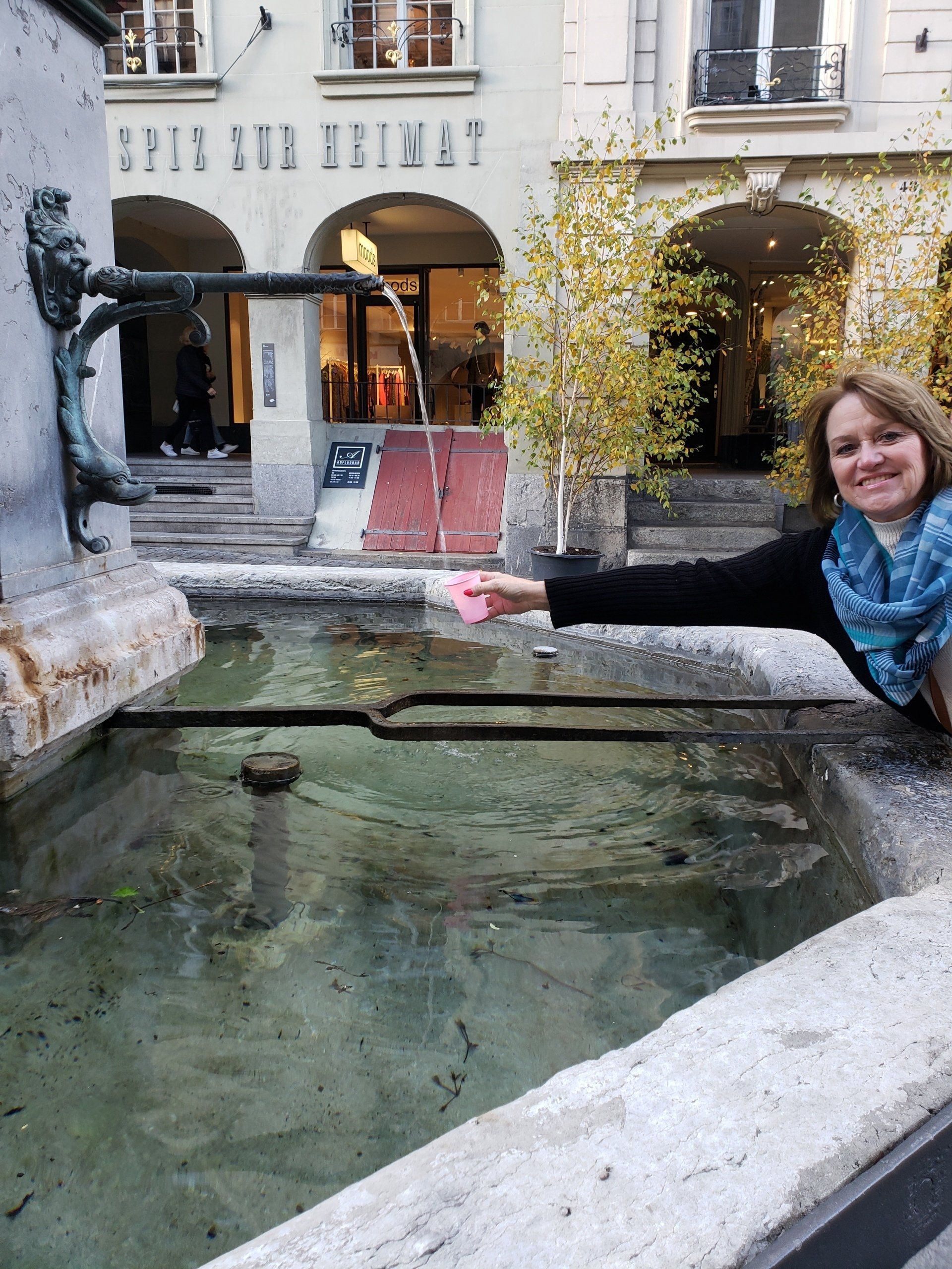 A woman is standing next to a fountain in front of a building with the word spitz on it
