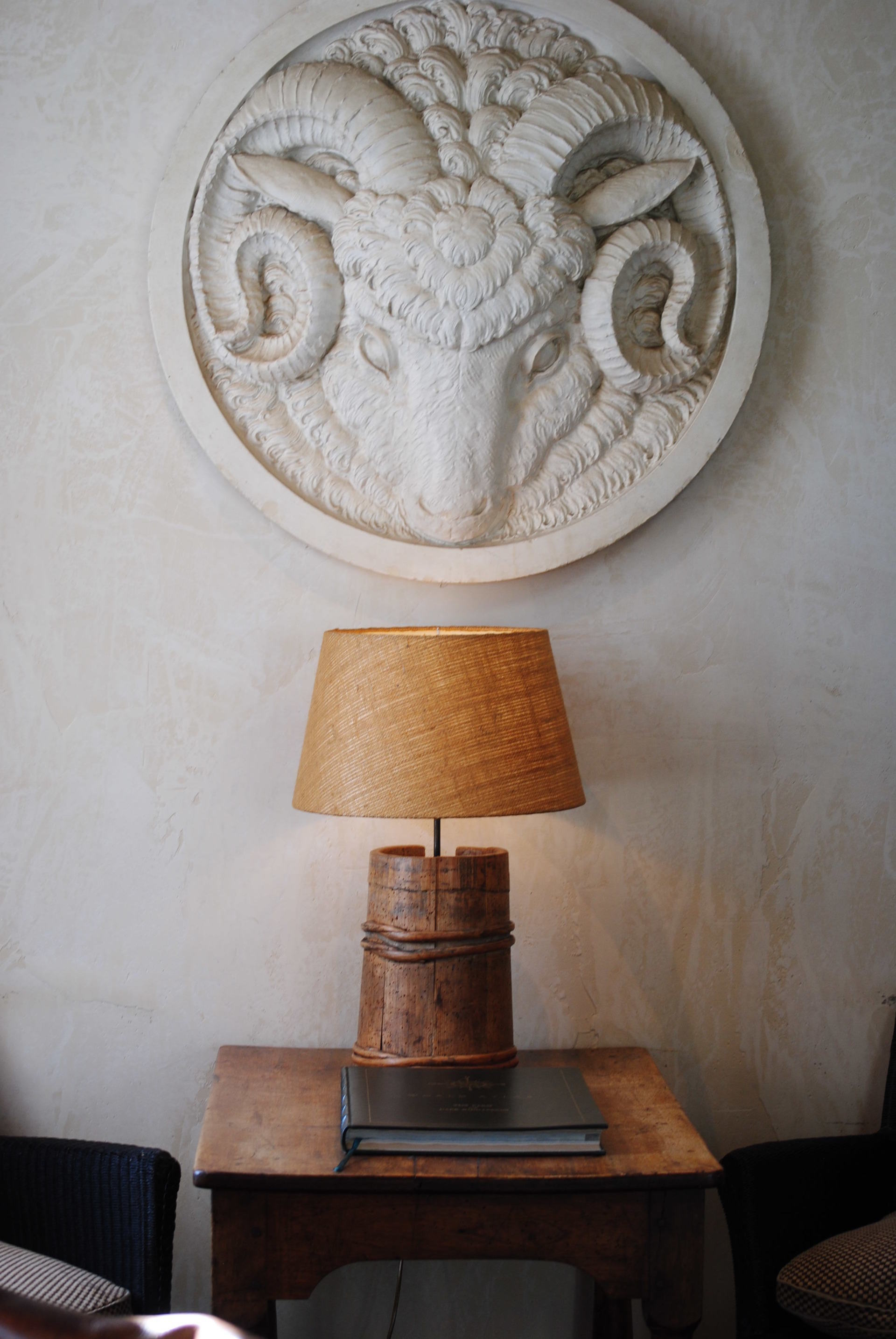 A lamp is sitting on a wooden table under a ram head plaque on the wall.