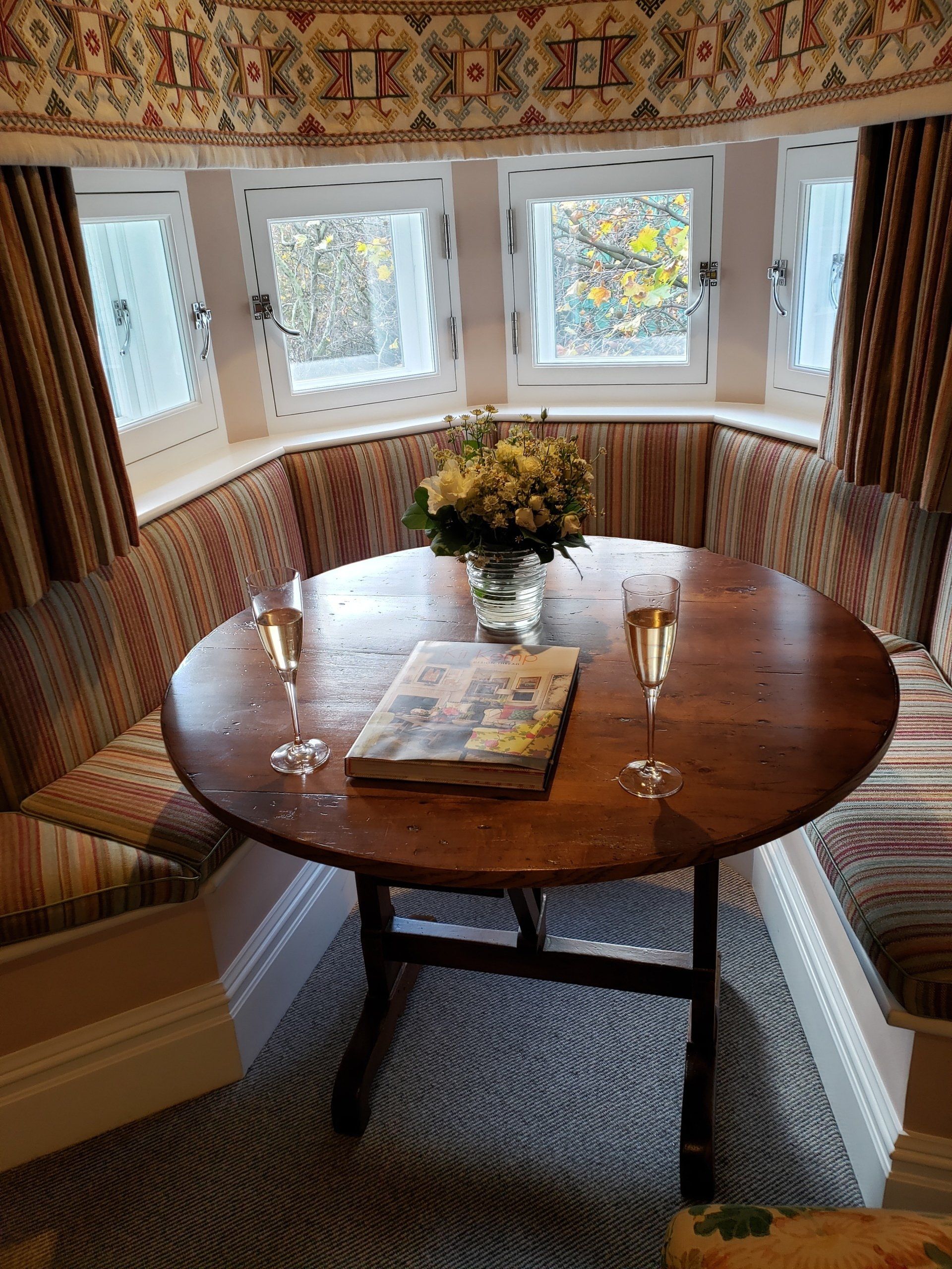 A round wooden table with two glasses of wine and a vase of flowers on it.