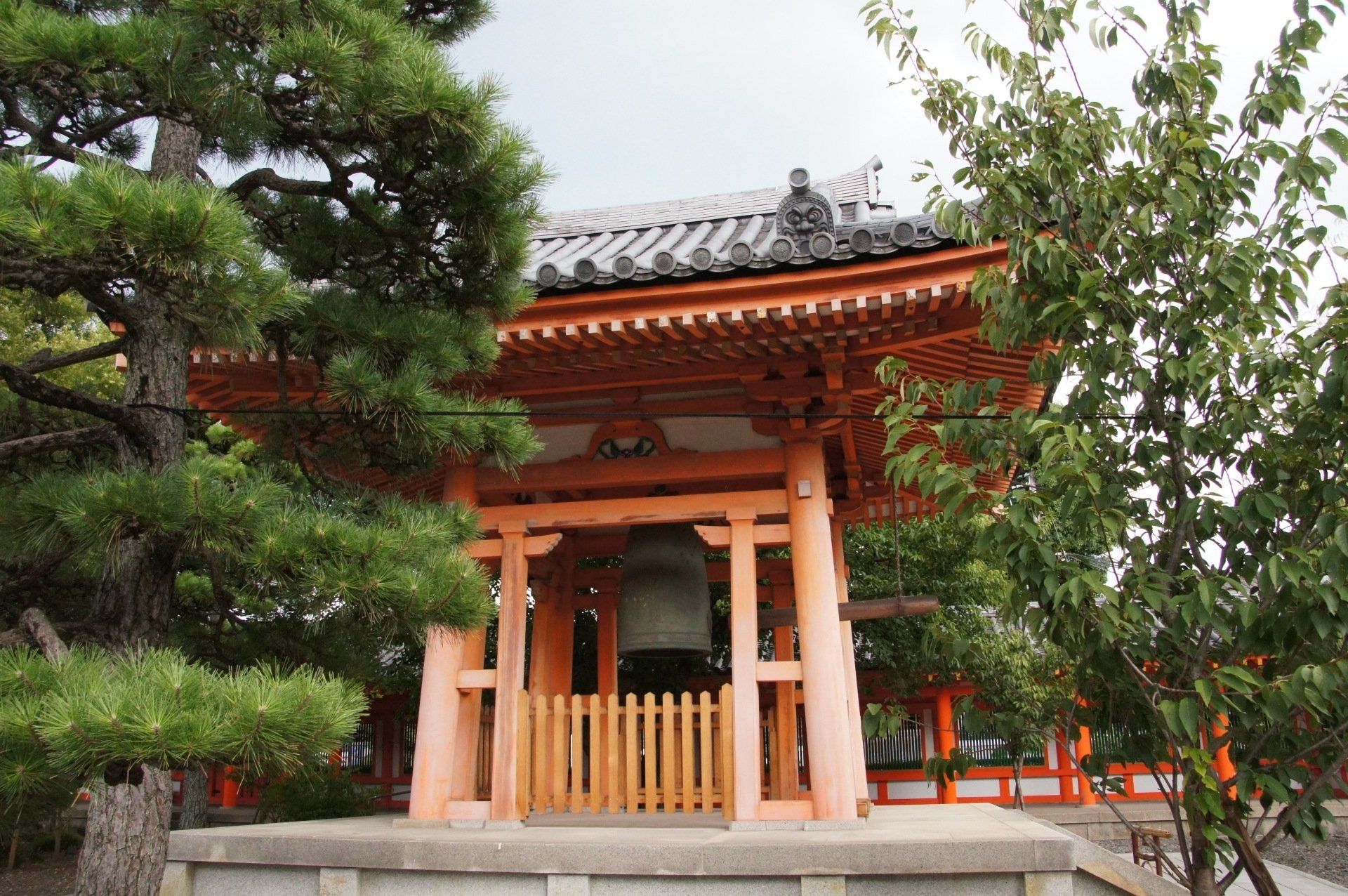 A large bell is hanging from the roof of a building surrounded by trees.