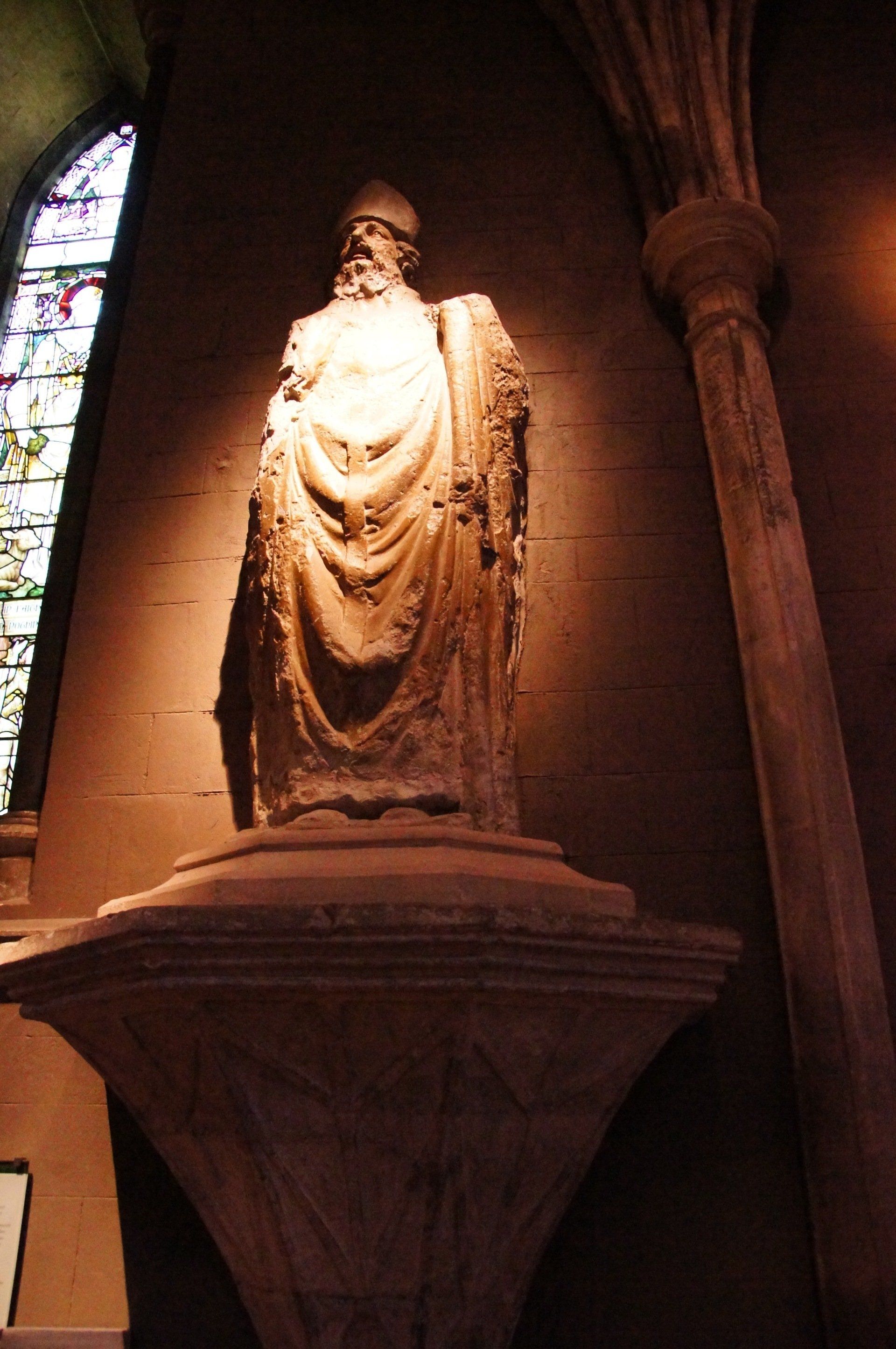 A statue of a man with a beard is on a pedestal in front of a stained glass window