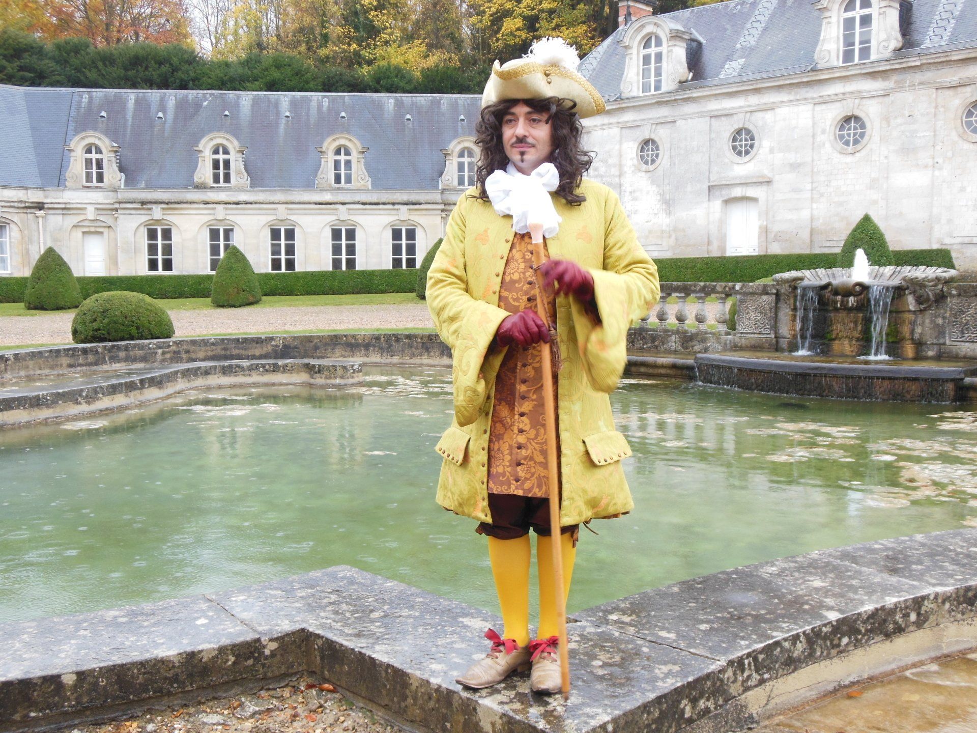 A man in a yellow coat and hat is standing in front of a fountain.