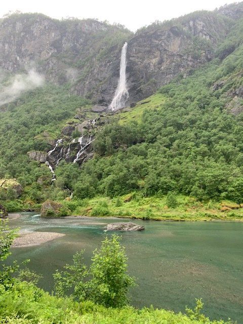 A waterfall is surrounded by mountains and trees next to a lake.