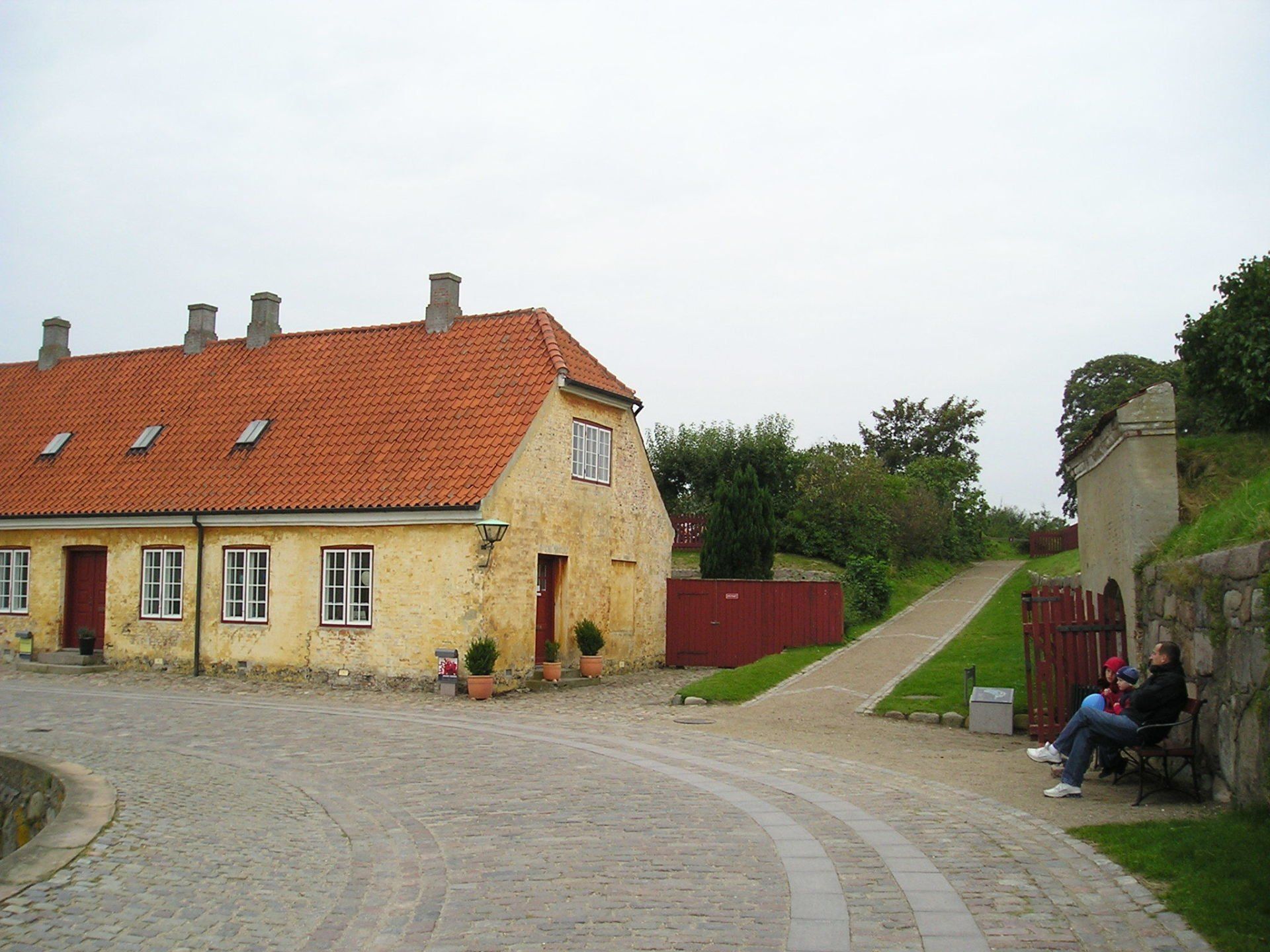 A man sits on a bench in front of a yellow building with a red roof
