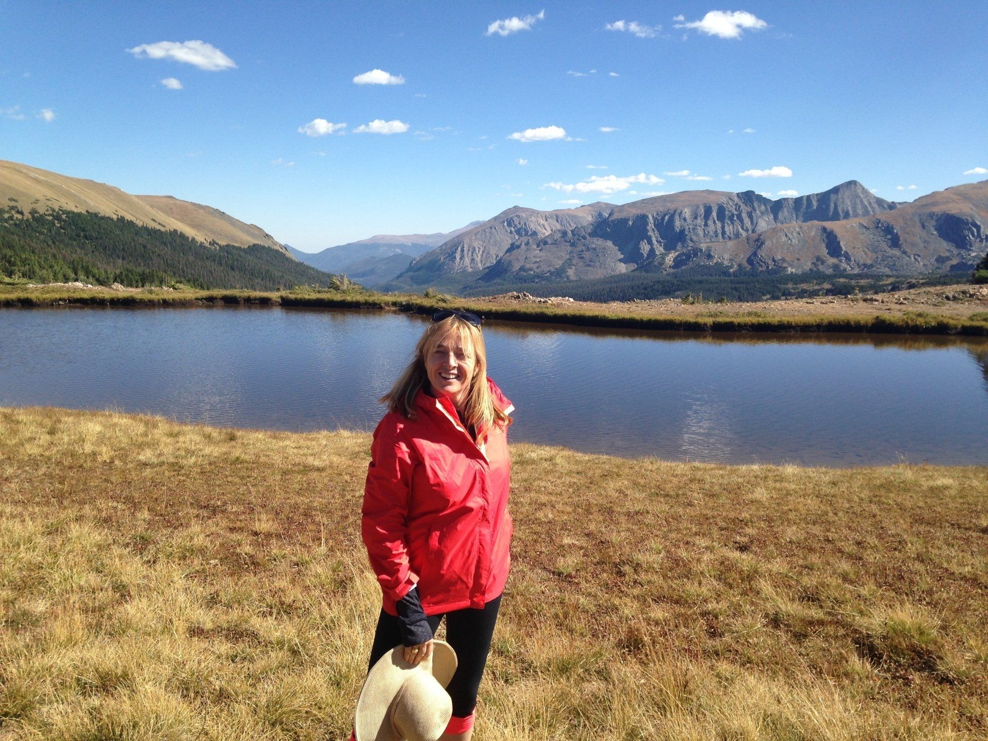 A woman in a red jacket is standing in front of a lake with mountains in the background.