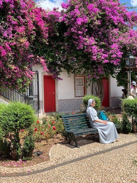 A woman is sitting on a bench in front of a building with purple flowers.