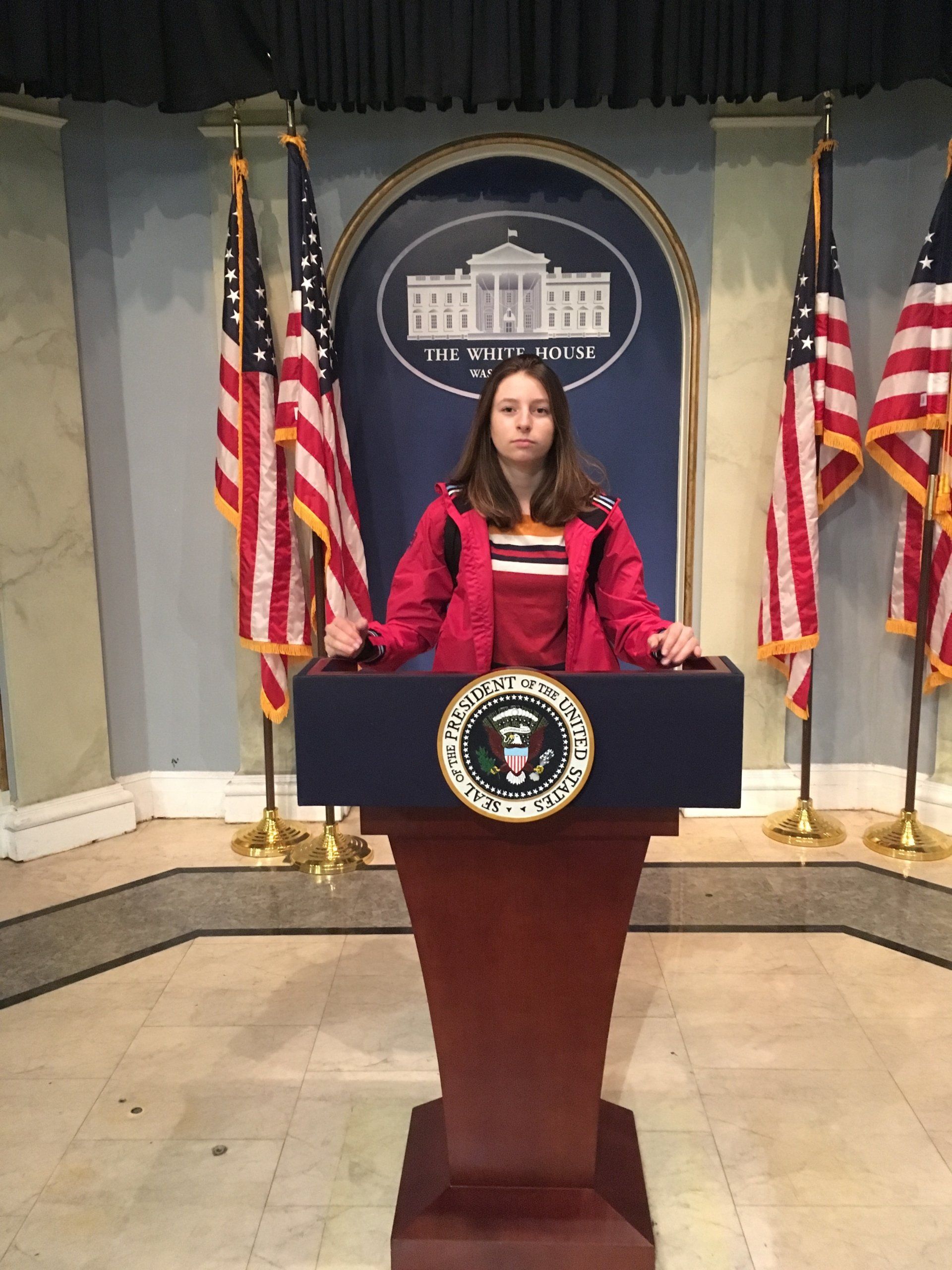 A girl in a red jacket stands at a podium in front of the white house