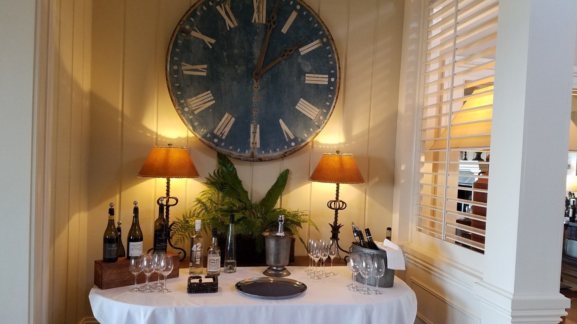 A large clock hangs above a table with wine bottles and glasses on it