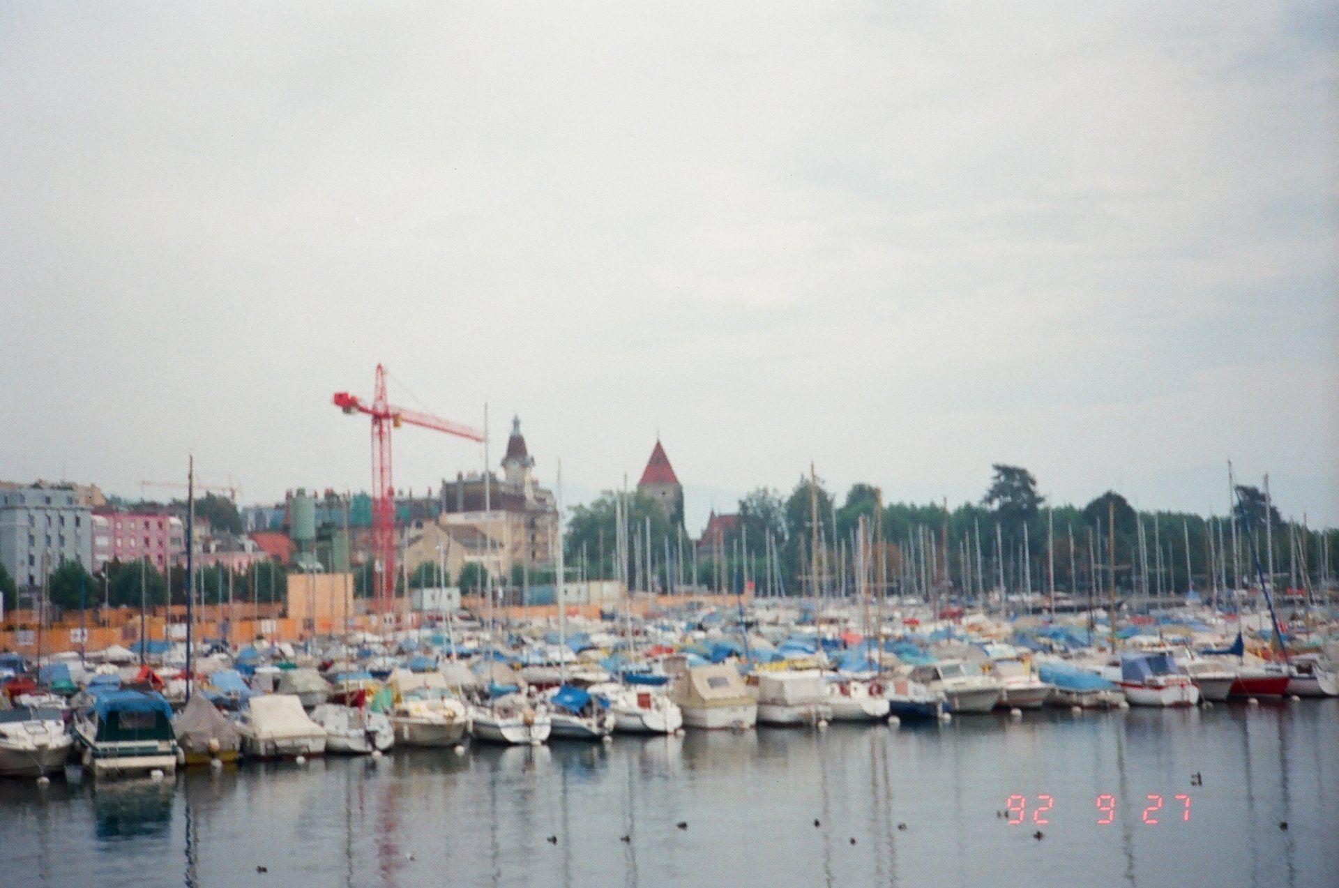 A lot of boats are docked in a harbor with a red crane in the background