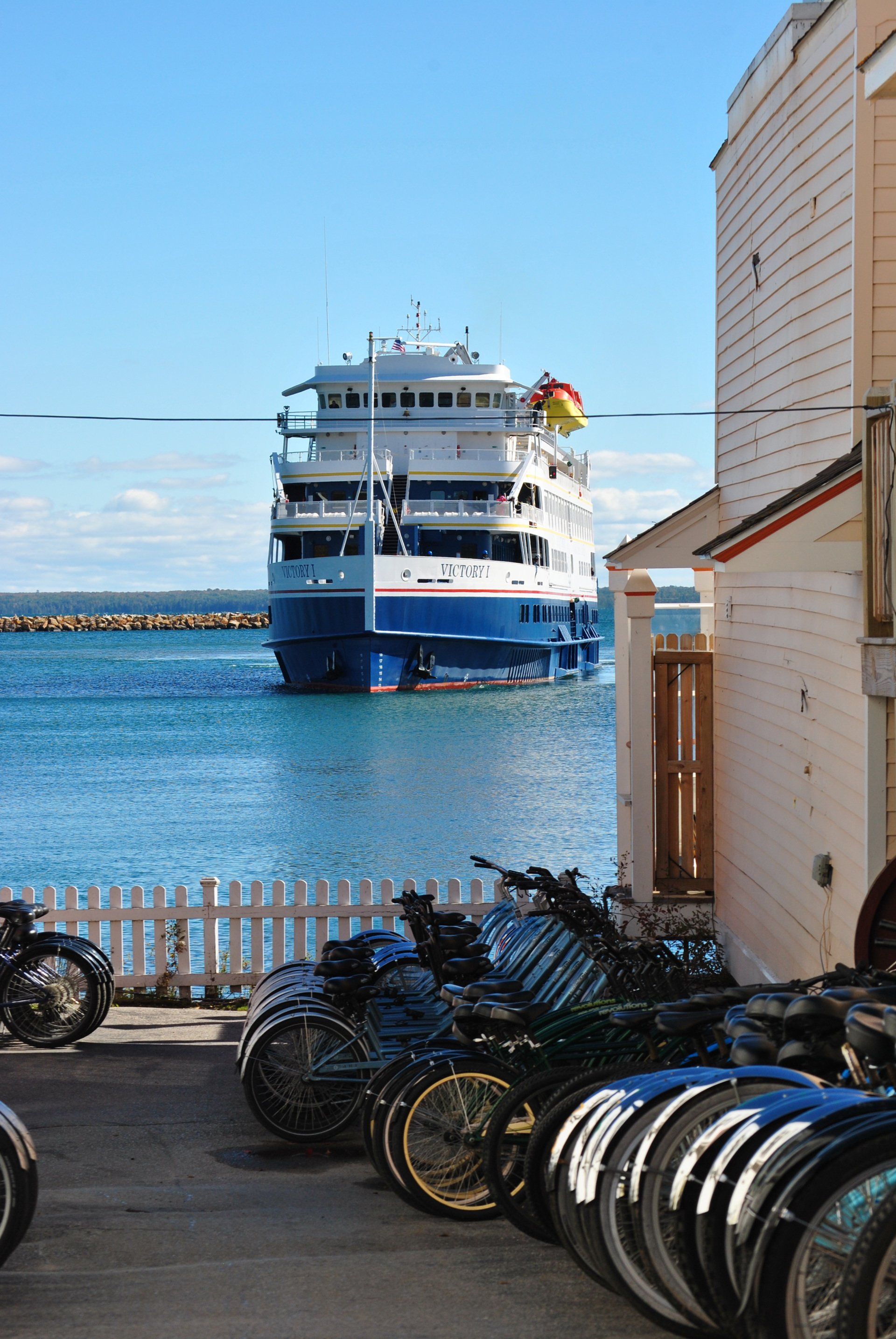 A row of bikes are parked in front of a building with a boat in the background