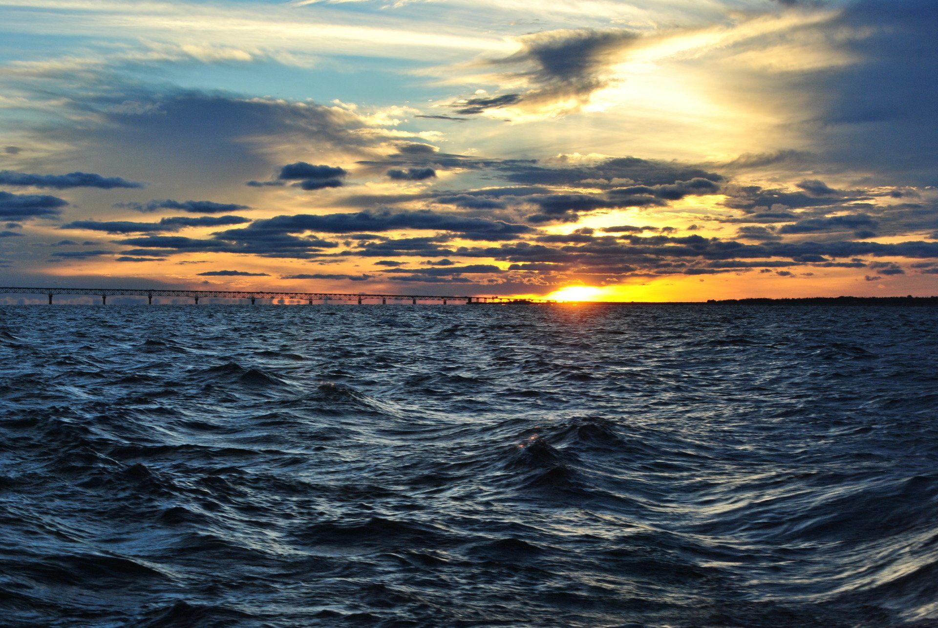 The sun is setting over the ocean with a bridge in the distance.