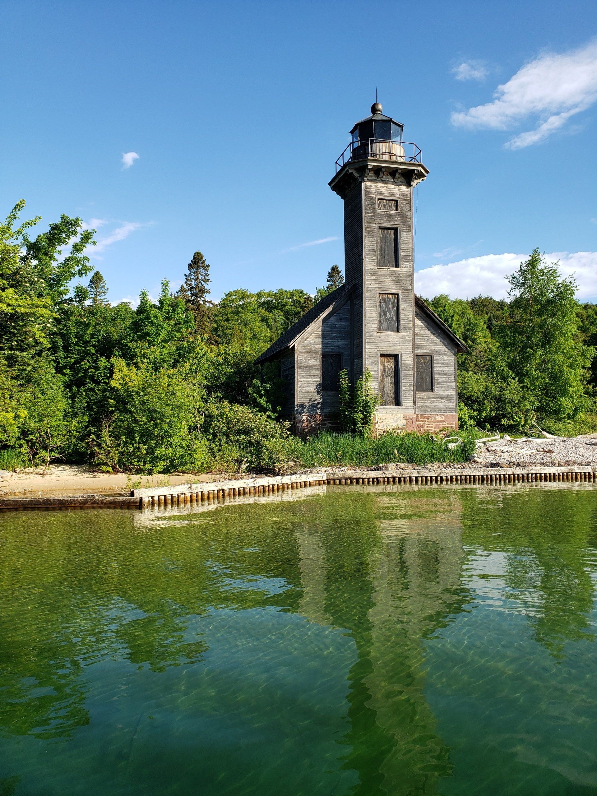 A lighthouse is reflected in the water of a lake surrounded by trees.