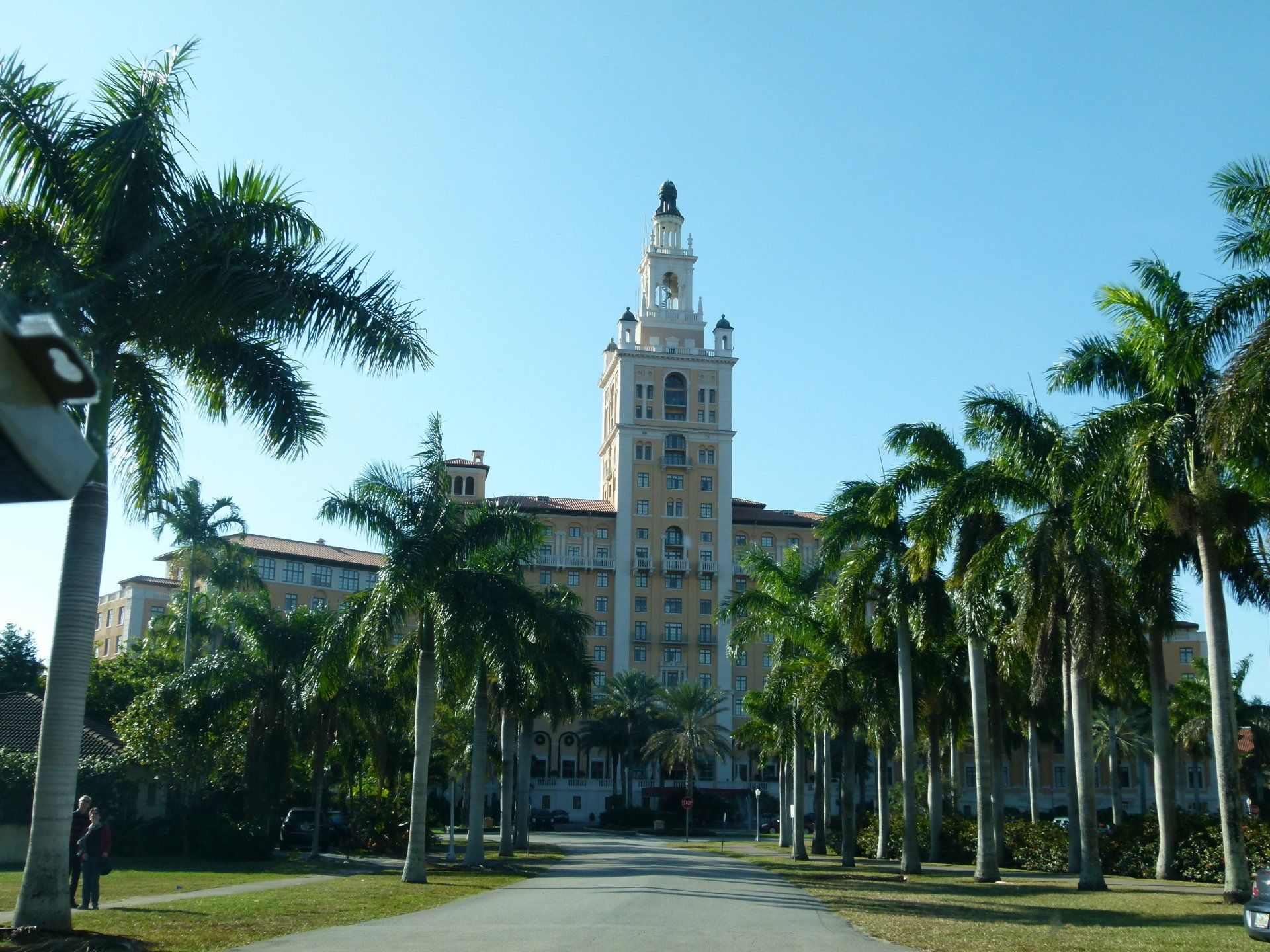 A large building with a clock tower is surrounded by palm trees