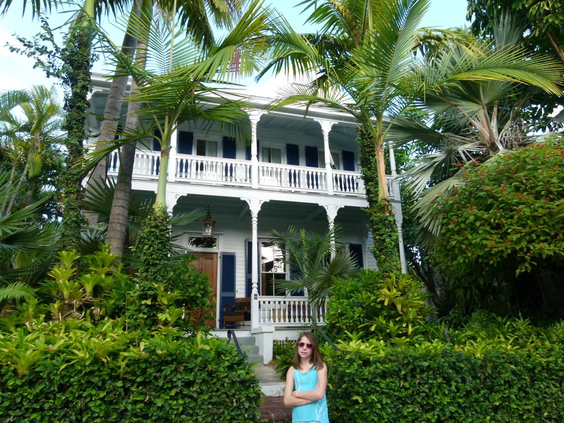 A woman stands in front of a large white house surrounded by palm trees