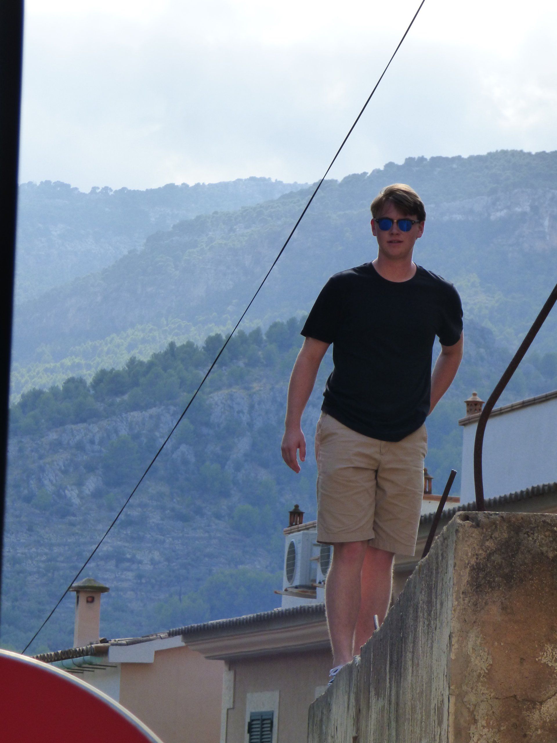 A man wearing sunglasses is standing on a wall with mountains in the background