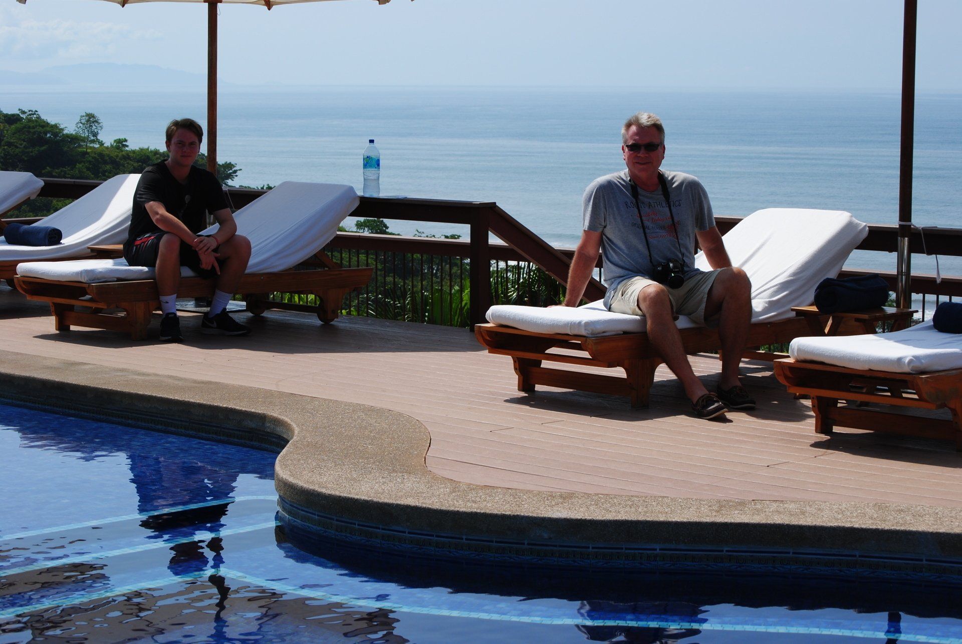 Two men sit on lounge chairs near a pool overlooking the ocean