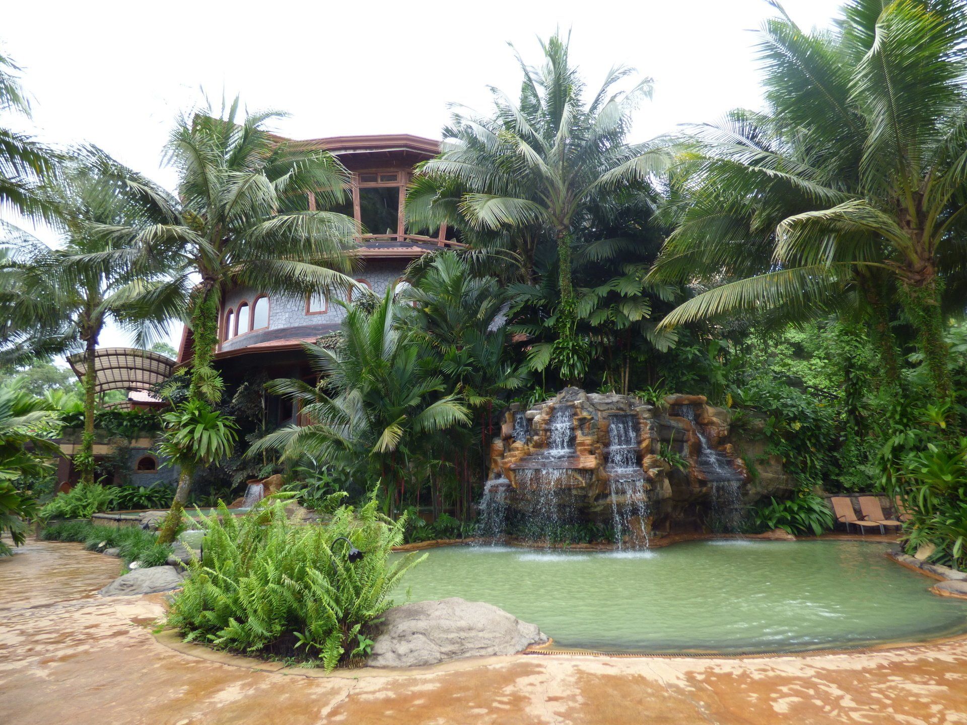 A waterfall is in front of a house surrounded by palm trees.