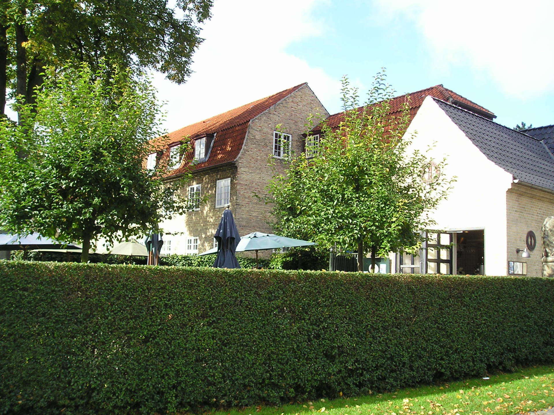 A large house with a red roof is behind a lush green hedge